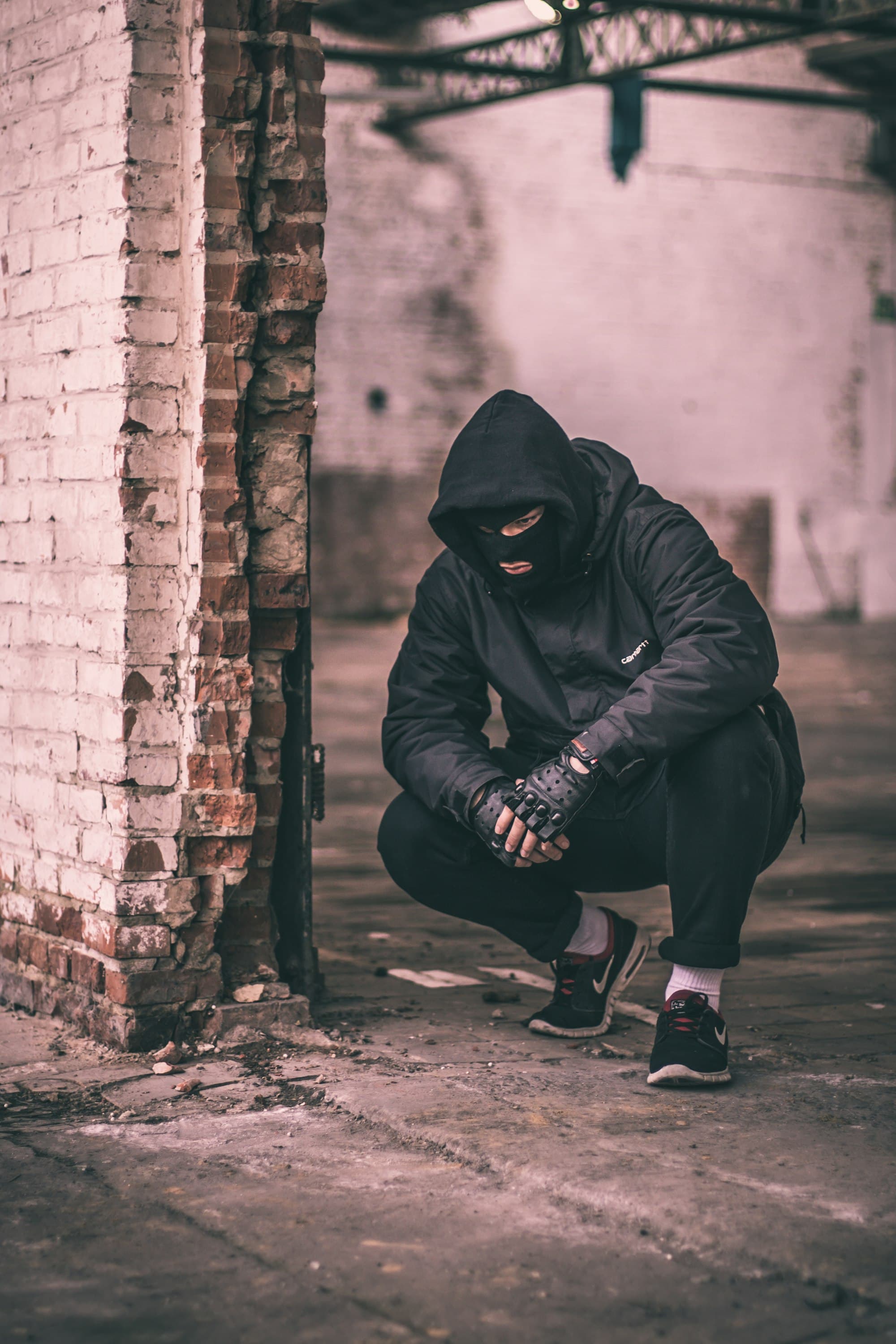 Man wearing dark clothes and a ski mask crouching near a wall.